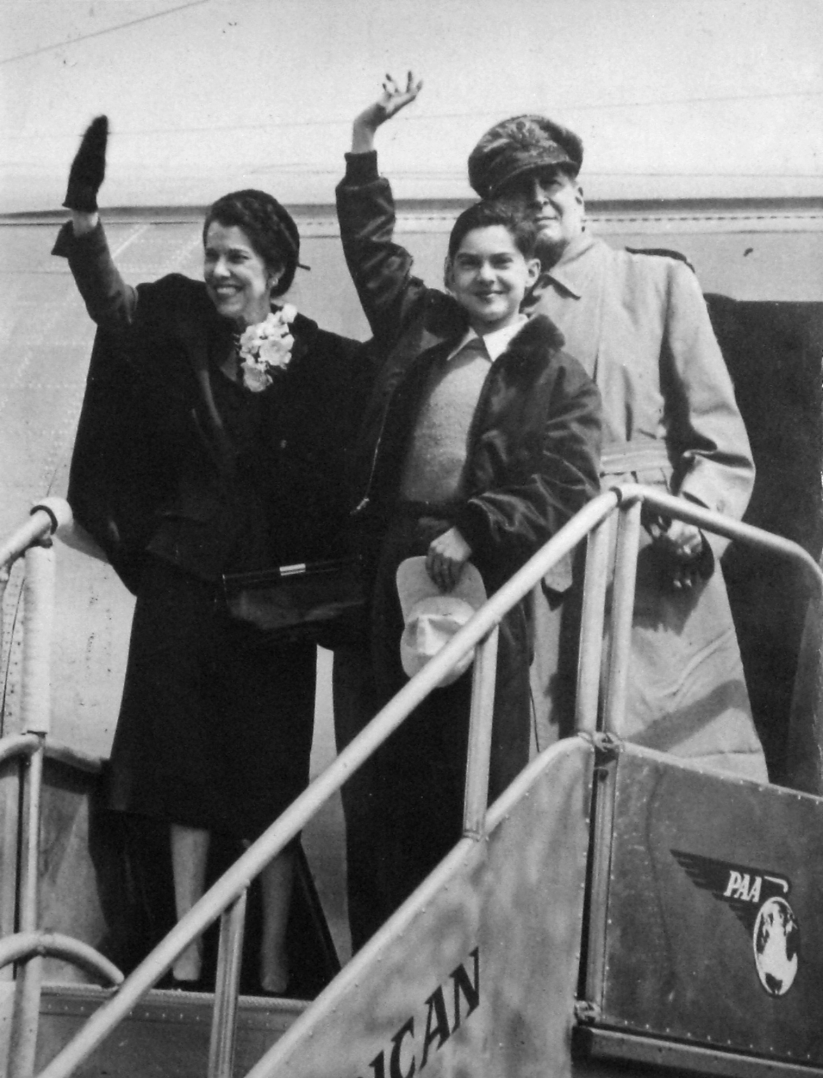 Black-and-white photograph of Douglas MacArthur with his wife and son, standing on the boarding ramp of an aircraft. Two of them are waving. 