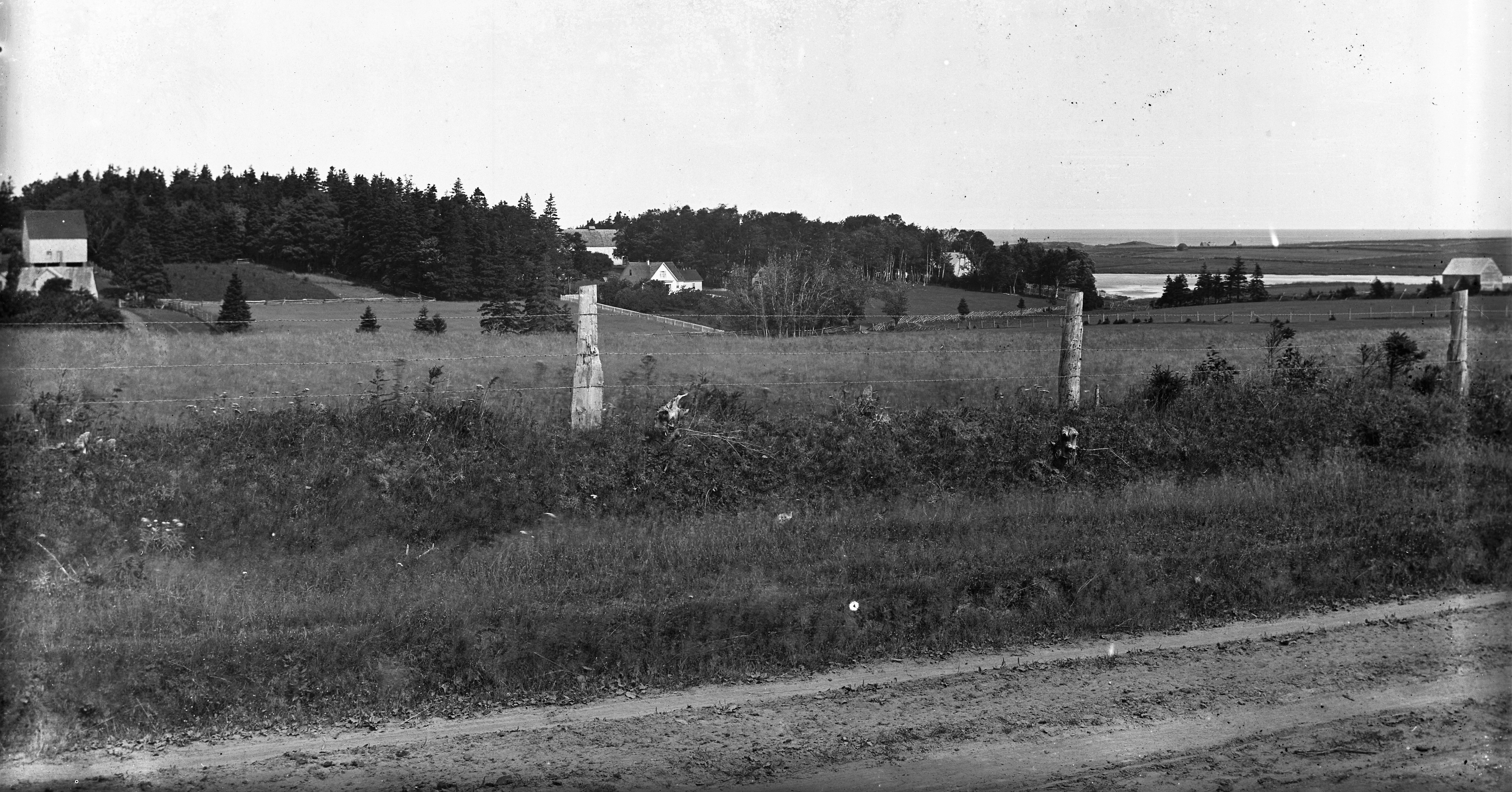 Black-and-white photograph of Cavendish. In the foreground is a fence in a field and in the background is a body of water behind farmland.