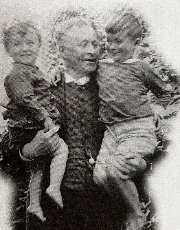 A cropped image of a smiling, grey-haired Ewen Macdonald holding his two young boys who are smiling and laughing. 