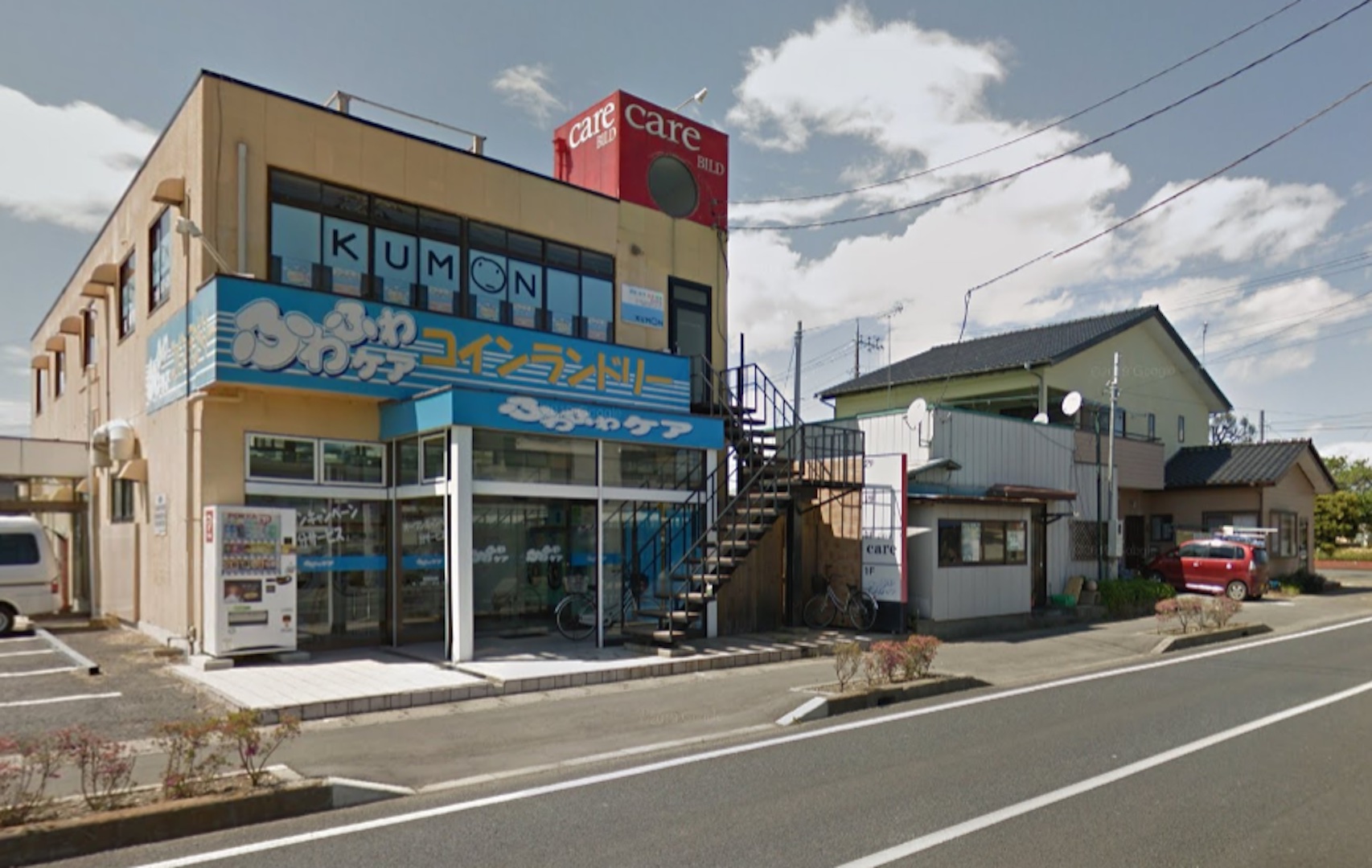 Photograph of Oarai, Ekimae Street, in 2013. A building with blue-and-yellow signage is situated across a street. There are other buildings to the left of it.