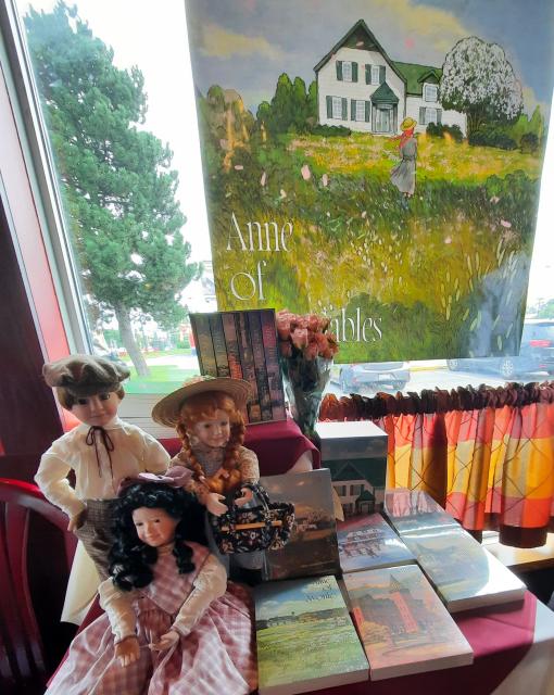 Display of books and dolls. Table display of three dolls and Thai book covers with poster of Anne of Green Gables behind.