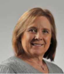 Photograph of Lesley Clement. She is a white woman with shoulder length blonde hair. She is facing forward against a gray background and is smiling.