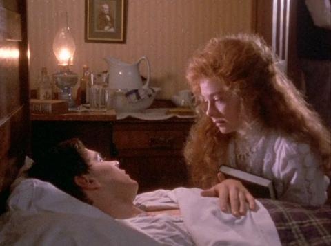 Photograph of a man with black hair lying in bed looking at a woman with red hair sitting at his bedside holding a book.  