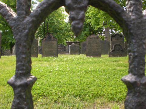 Photograph of a graveyard taken from near ground level. There is an iron arch that acts as a blurred border that the graves are focused through. 