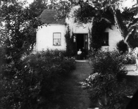 Black-and-white photograph of house with two windows on either side of a door and two women sitting on doorstep. The house is surrounded by flowering plants and trees. 