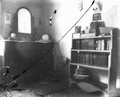 Lucy Maud Montgomery's room downstairs, ca.1900's. Cavendish, P.E.I. Black-and-white photograph of a room with a vanity on the left and a bookcase on the right. There are several crack lines in the bottom left-hand corner of the image. Two of the cracks continue across the image.