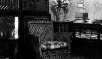 Black-and-white photograph of a chair in the centre of the image and a bookcase to the right. In the back on the left is an enclosed bookcase. 