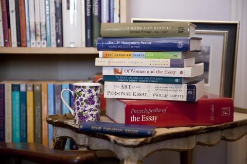 Collection of Essays and Anthologies Photograph of a stack of books on an antique table. A teacup with purple flowers is sitting on the table beside the books. There is a filled bookcase in the background.